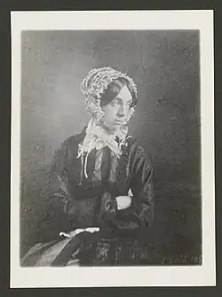 Valentine Delessert in a dark dress and white bonnet sitting with arms crossed staring away