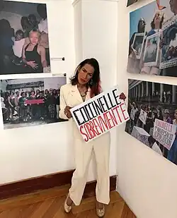 Woman in pantsuit holds a sign reading "Coccinelle Sobreviviente"