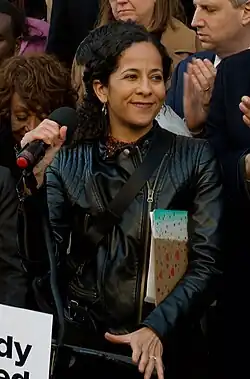 A woman grabs a microphone at a lectern, looking to her right. Under one arm is a teal-painted clipboard.