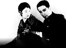 A black and white movie still showing a man and a woman clasping hands. The woman is wearing a kimono and is looking over to the man who stares straight ahead.