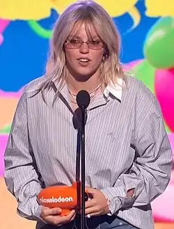 A white woman with blonde hair and bangs holds an orange award and speaks in a microphone