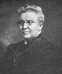 Black and white photo of a white-haired woman wearing glasses, and dark, high-collared clothes