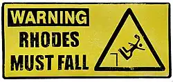 A Road sign saying Rhodes Must Fall, showing a triangle next to it with a man falling from a pedestal.