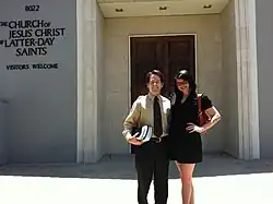 Man with light skin and dark hair and woman with dark hair and light skin stand in front of a building with a sign that reads "The Church of Jesus Christ of Latter-day Saints; Visitors Welcome"