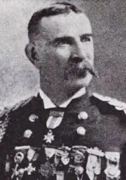 Head and shoulders of a white man with a drooping mustache, wearing a military jacket with shoulder boards, one medal at the neck, and a row of medals across the entire chest.