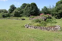 Low stone walls in grass, surrounded by trees with a house in the distance.