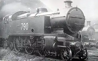 A side-and-front view of a large 2-6-4 steam locomotive at a locomotive depot. It is a tank locomotive with large rectangular water tanks either side of the boiler; they stretch from above the front driving wheels back to the cab. Members of the driving crew can be seen in the cab.