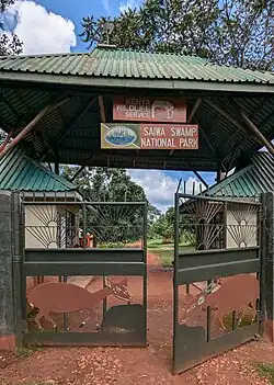 Dark green metal gates decorated with metal art of Sitatunga, with green metal roof over the entryway, a brown sign which reads "Kenya Wildlife Service" with the KWS logo, and another brown sign which reads "Saiwa Swamp National Park" and includes an illustration of swamp vegetation