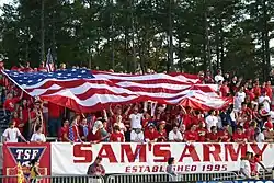 Soccer fans, dressed in red, cheer in bleachers as they hold a large American flag over themselves at a soccer match.