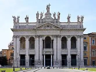 Ornate facade of the basilica with columns, main door, and statues of the twelve Apostles on the roofline, with a Latin inscription below them