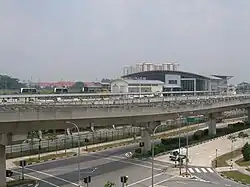 An elevated view of the Sengkang Depot building, a modern, large structure with a curved roof, situated next to an elevated LRT track. Some trains were parked on the top level where the LRT trains are tabled. Below the tracks are roads with traffic lights and some vehicles.