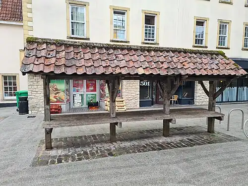 Colour photograph of the restored Shambles in the Market Place at Shepton Mallet. The bench and curved roof trusses are made of oak, and the floor is made of local stone and is brown in colour. The roof is constructed from red pantiles. Modern shops can be seen in the background.