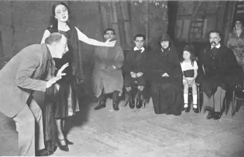 stage scene with director bending forwards, actress standing gesticulating and small family sitting in row