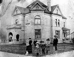 Black and white photo of the Smoot family in front of their home in Provo, Utah. The family is dressed in typical turn-of-the-century attire. Reed and his wife, Allie, are seen with five of their children, including one baby in Allie's arms. A medium-sized dog is sitting in front of the family.