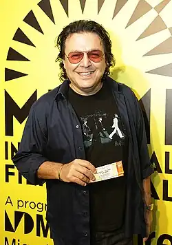 A man is wearing sunglasses and staring at a camera while holding tickets to an event.