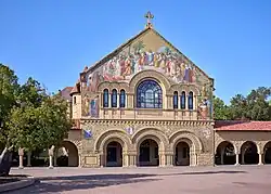 Floodlit view at night of the façade of the church. The facade has two stories, At the lower level are three arched doorways. In the upper level is a large central arched window flanked by tall narrow triplet windows. Above the windows rises a gable, richly decorated with a mosaic of a biblical theme and surmounted by a cross.