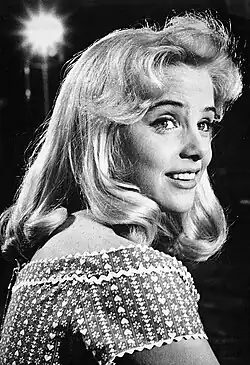 Close-up black-and-white portrait photo of a smiling young woman with long blonde hair in a studio, brightly illuminated by set lights
