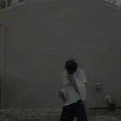 An unsaturated photograph of a person standing in front of a house with their head down.