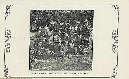 An old black and white photograph of a group of about 20 people posing for a photo in front of some trees.