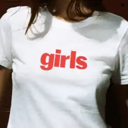 A photograph of a woman from the neck down wearing a white T-shirt with the word "girls" on it in red text.