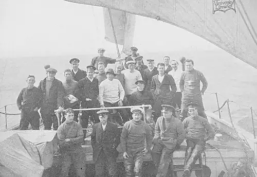 A group of men on board a ship, identified by a caption as "The Weddell Sea Party". They are dressed in various fashions, mostly with jerseys and peaked or other hats. The rough sea in the background suggests they are sailing into stormy weather.