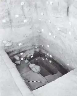 A rectangular hole in the ground, with high walls forming two sides opposite the camera. There are labels denoting different layers and features on the vertical faces, and inside the hole the ground is stepped as different parts have been excavated to different depths.