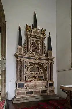 This image shows a large, elaborate multi-tiered wall monument in a church setting. The monument features a central arched section supported by dark columns, within which lies a single reclining effigy figure carved in light-coloured stone on a tomb chest, with an inscription panel above. The structure is topped by an ornate heraldic display showing multiple rows of coat of arms surrounding a central panel, flanked by four tall dark obelisks with golden finials. Two golden winged creatures crown the composition on either side of the tallest central spire. The monument combines dark and light stone materials with gilded decorative elements throughout, creating an imposing vertical structure that dominates the white church wall on which it is mounted.