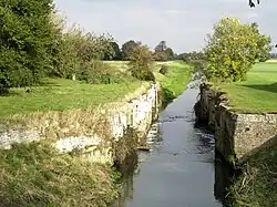 The best preserved of the lock chambers at Alvingham, the retaining walls of brick and stone are intact, but there are no gates. We are looking downstream, away from the weir formed by the upstream sill, which is not visible. On either side of the channel are flat green meadows, and framing those to the edges of the picture are dense woodland. It is a bright sunny day and the trees and grass are a bright green. The data for the picture say it was taken in October 2007, but the vivid green of most of the trees, and particularly one just on the right of the lock, looks like spring.