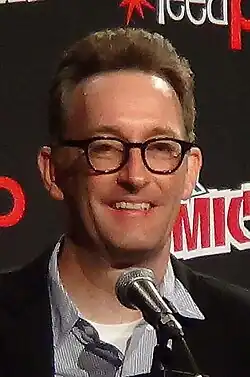 Tom Kenny, a tall white man with brown, curly hair and glasses, seats at a microphone