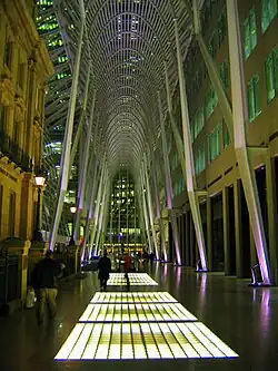 A towering metal gridwork catenary arch forms a gallery over a shiny concrete concourse inset with glowing grids of glass. Beyond the arch, city lights of tall buildings are visible. A four-story historic stone building stands to the left; a plain modern building rises out of sight to the right.