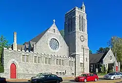 Two pointed gray stone buildings seen from across a street, with cars passing in front and a traffic light visible from the center to the upper left. The one on the left has a tall square stone tower with a smaller tower topped by a cross at the top. The one on the left has a large round window in the middle and a cross at the point on the front. Both have open red doors.