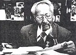 An elderly mustachioed Asian man with oval glasses reading at a desk