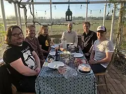 Six white men in casual clothing sitting around a table in a ground-level sunroom. Plates, glasses, drinks, and opened food containers are sprawled across the table.
