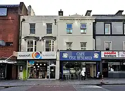 Four terraced buildings, three of them small, all two storeys, in a high street setting. The two central buildings are white; the building on the furthest left is of red brick, and the building furthest right is grey.