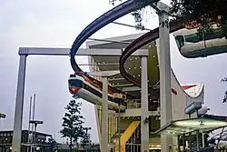 A tall, white building with a curved roof serves as a monorail station. A white train leaves the station while another arrives.