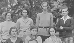 Four women stand behind three seated women, all facing the camera.