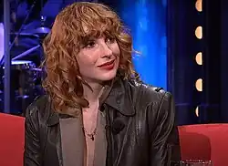 Vica Kerekes wearing a dark brown leather jacket over a light brown, low-cut top with lapels, sitting on a red couch, grinning slightly, and looking left of camera