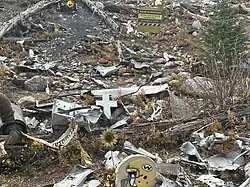 A hillside is scattered with airplane wreckage and personal tributes at the Wichita State University football team plane crash site. A yellow football helmet, a sunflower, and a religious statue are visible near the debris. A sign reads, “Riding to Remember Wichita State University Football Team, October 2, 1970,” and a yellow jersey hangs from a tree trunk. The area is surrounded by trees and rocky terrain.