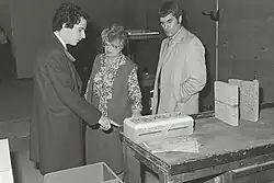 Monochrome photograph of William Waldegrave's visit to the heat laboratory at the University of Salford in 1981. Waldegrave is shown stood with Stuckes, the then head of the laboratory, and John Ashworth. They are photographed behind a table holding anechoic poly blocks.