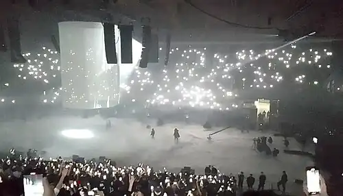 Crowds watch shawdowy figures on a foggy stage. Members of the crowd are holding the phone flashlights in the air.