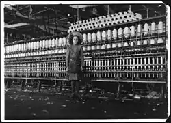 A black and white photograph of a young girl standing in front of mill machinery