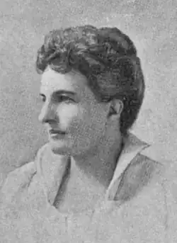 A white woman with dark wavy hair in an updo, wearing a light-colored top with a standing collar