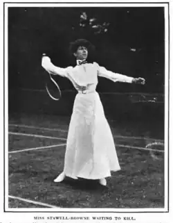A white woman standing on a tennis court in tennis whites, including an ankle-length skirt, holding a tennis racquet behind her head in a position of readiness